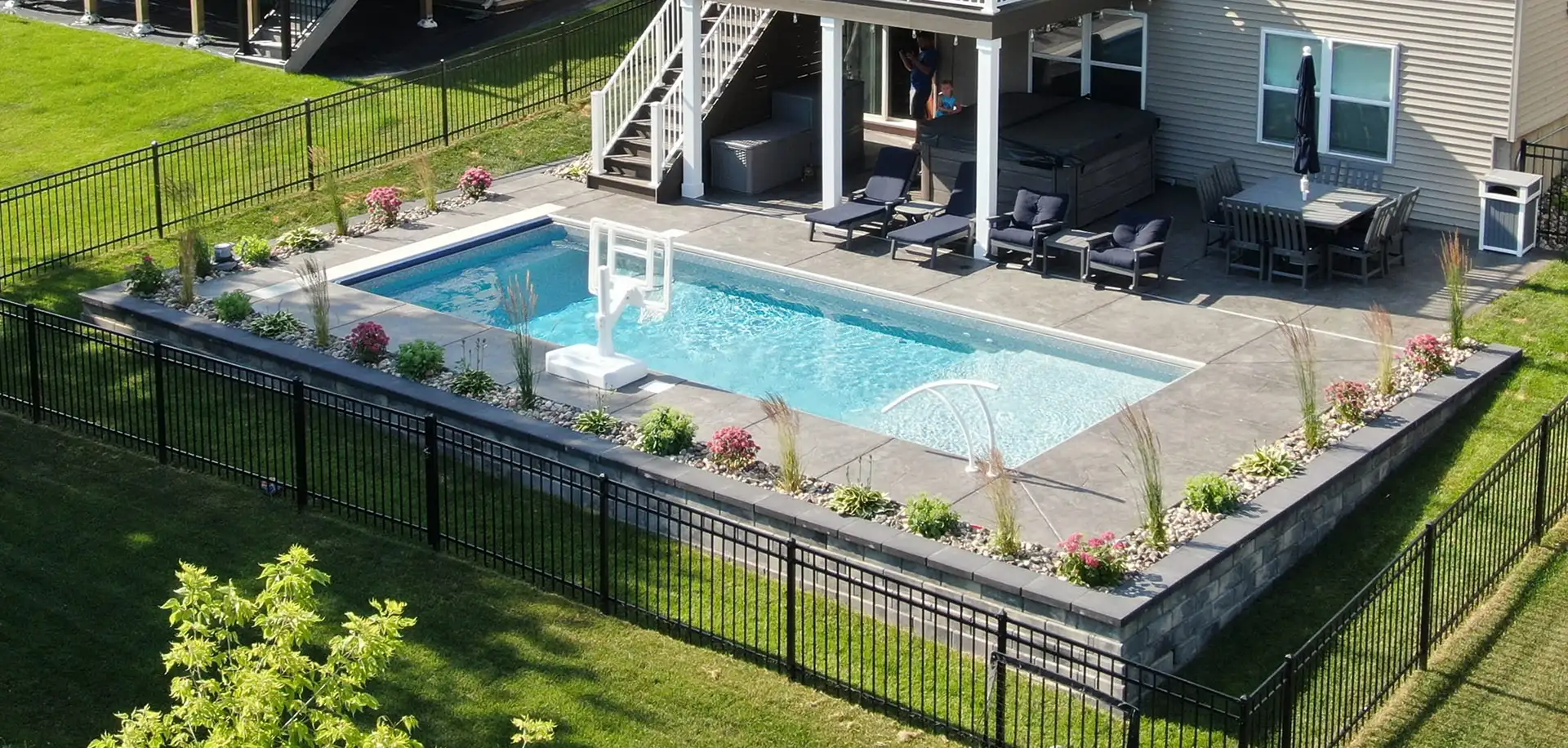 Backyard swimming pool enclosed by a black metal safety fence with a self-closing gate around a concrete pool deck. The fenced pool area beside a house patio illustrates practical pool safety tips for keeping children and pets away from the water.