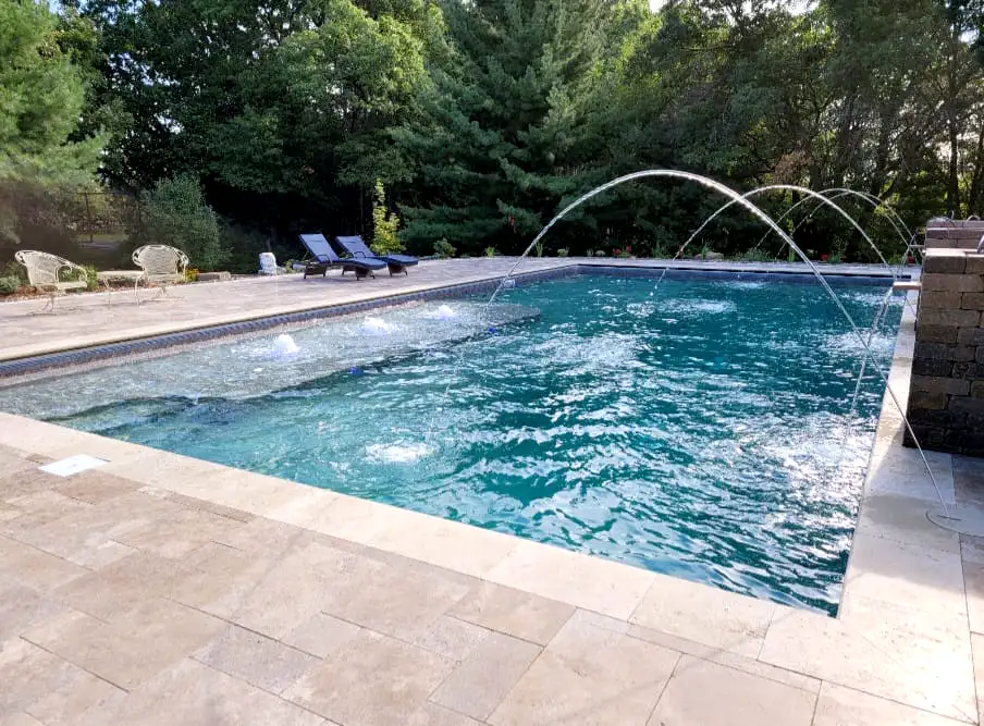 Outdoor rectangular swimming pool with clear blue water, bubbling jets, and three arcing water features spraying into the pool, surrounded by light stone pavers, lounge chairs, and trees in the background.