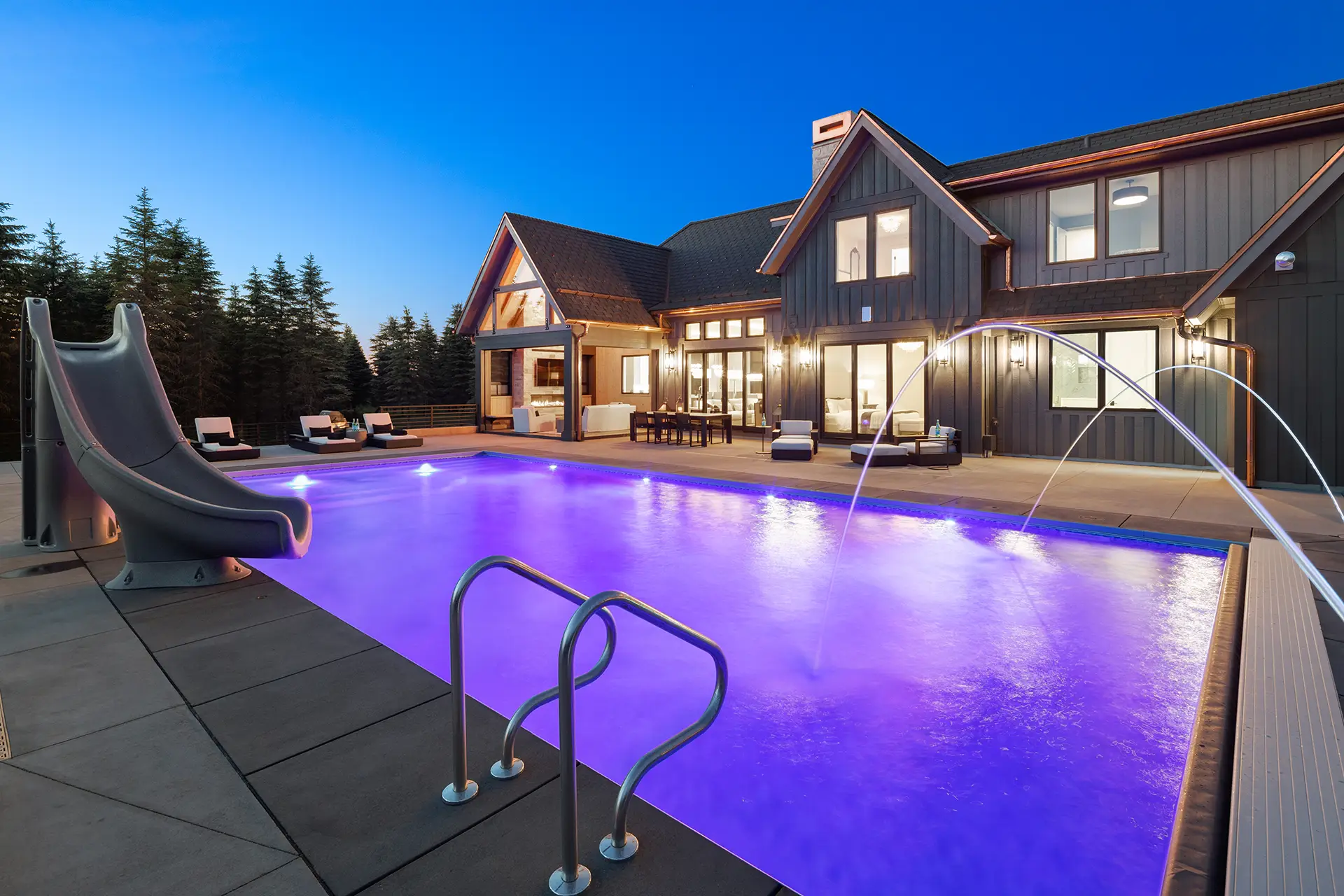 Luxury home in Bayport, MN, with a glowing purple-lit in-ground pool at dusk, featuring a water slide, deck jets, and poolside lounge chairs. The two-story house has large windows, warm exterior lighting, and is surrounded by tall evergreen trees, creating a serene and upscale outdoor setting.