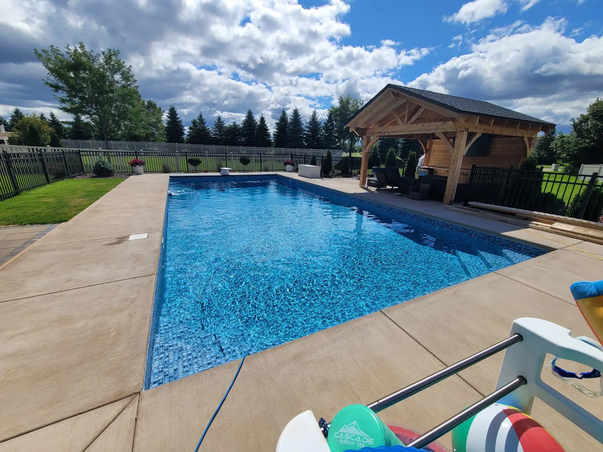 A backyard swimming pool in Lakeville, MN, with sparkling blue water, is surrounded by a concrete deck, black iron fence, and neatly manicured lawn.