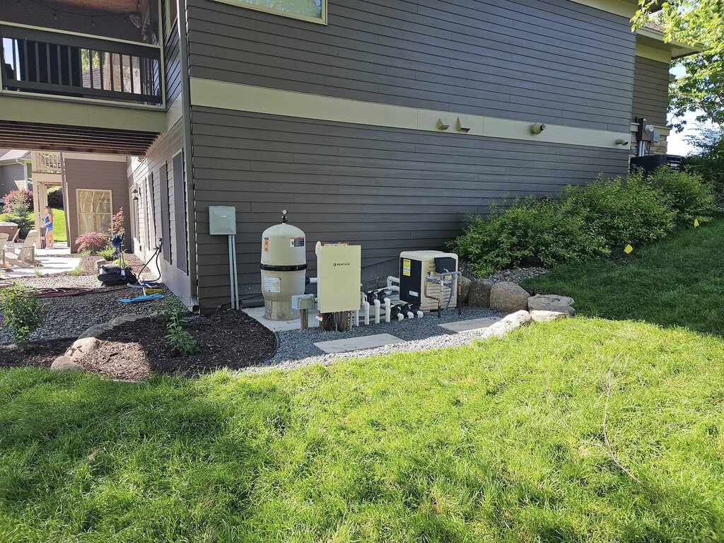 Pool equipment pad beside a house, with a filter tank, Pentair control panel, heater, and pump plumbing on a gravel bed.
