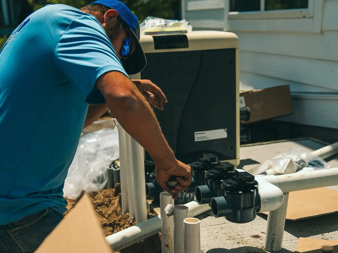 Pool technician wearing a blue shirt and cap adjusts valves and PVC plumbing on a pool filtration system beside a house during routine pool maintenance.