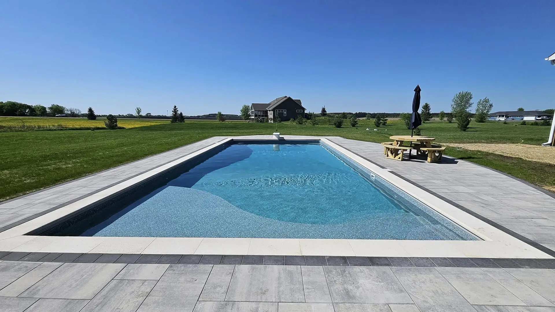 A saltwater swimming pool with sparkling blue water sits in a spacious Dellwood, MN backyard surrounded by green grass and open fields.