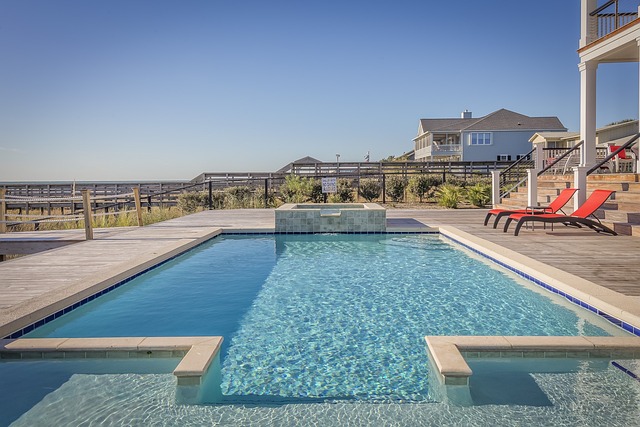 In-ground swimming pool with a raised spa, surrounded by a wooden deck with red lounge chairs, under a clear blue sky.